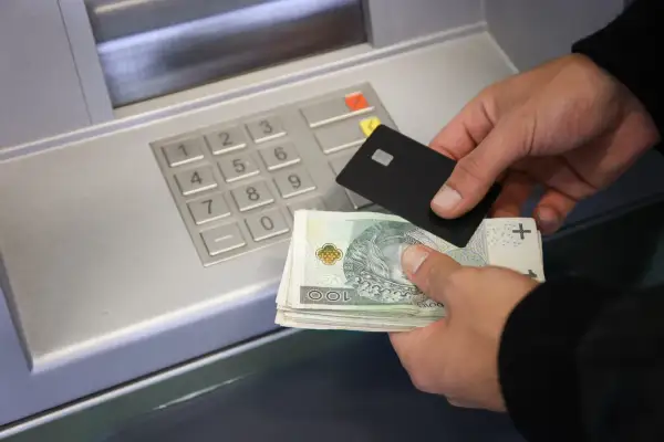 Ręce trzymające banknoty i kartę zbliżają się do bankomatu, pokazując, jak wpłacić pieniądze na konto.